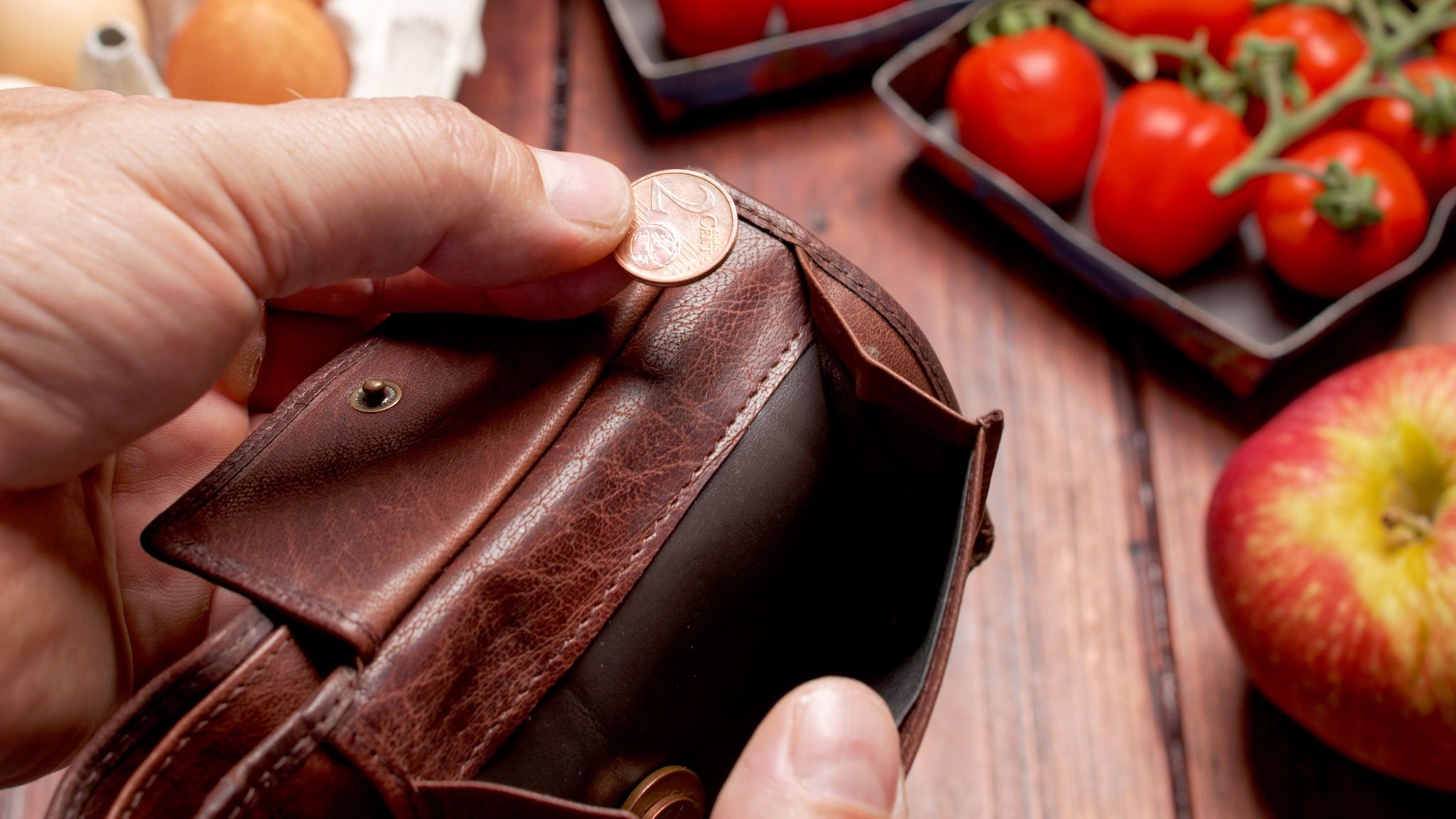 Hand hält eine leere Geldbörse und eine 2-Cent-Münze, im Hintergrund ein Apfel, Paradeiser und Eier auf einem Tisch