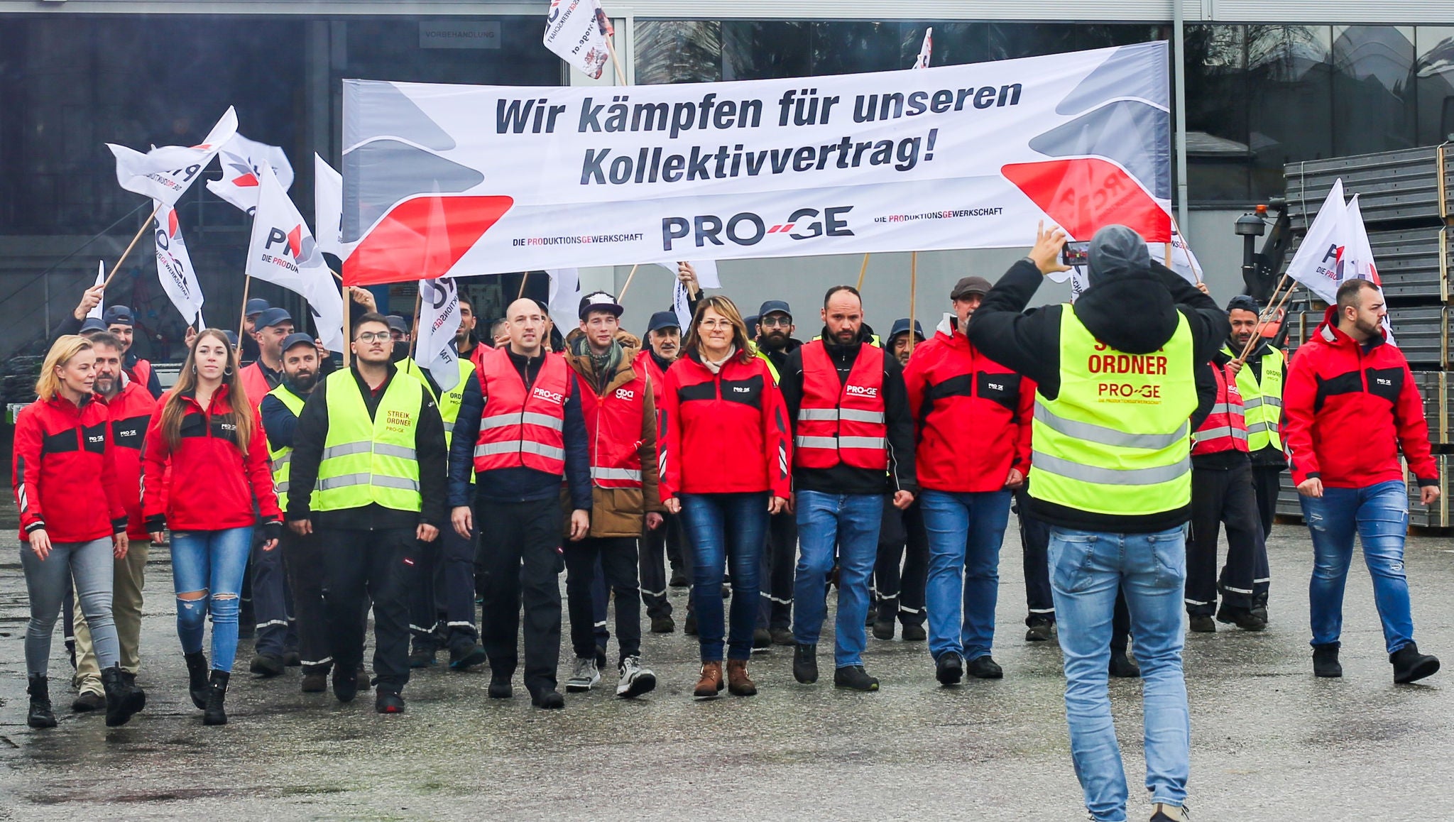Beschäftigte mit Warnwesten, Fahnen und einem Transparent mit der Aufschrift "Wir kämpfen für unseren Kollektivvertrag" ziehen vor das Werkstor von Collini in Salzburg.