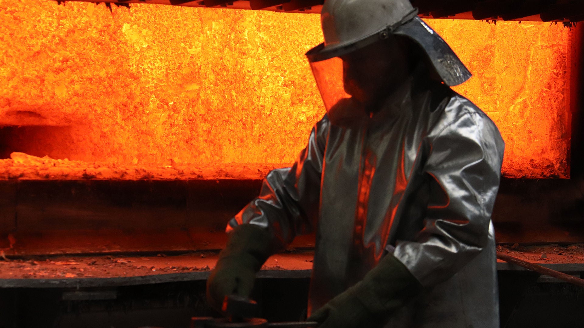 Im Hintergrund der Hochofen mit orange-gelbem Feuer, im Vordergrund eine dunkle Arbeiter-Silhouette