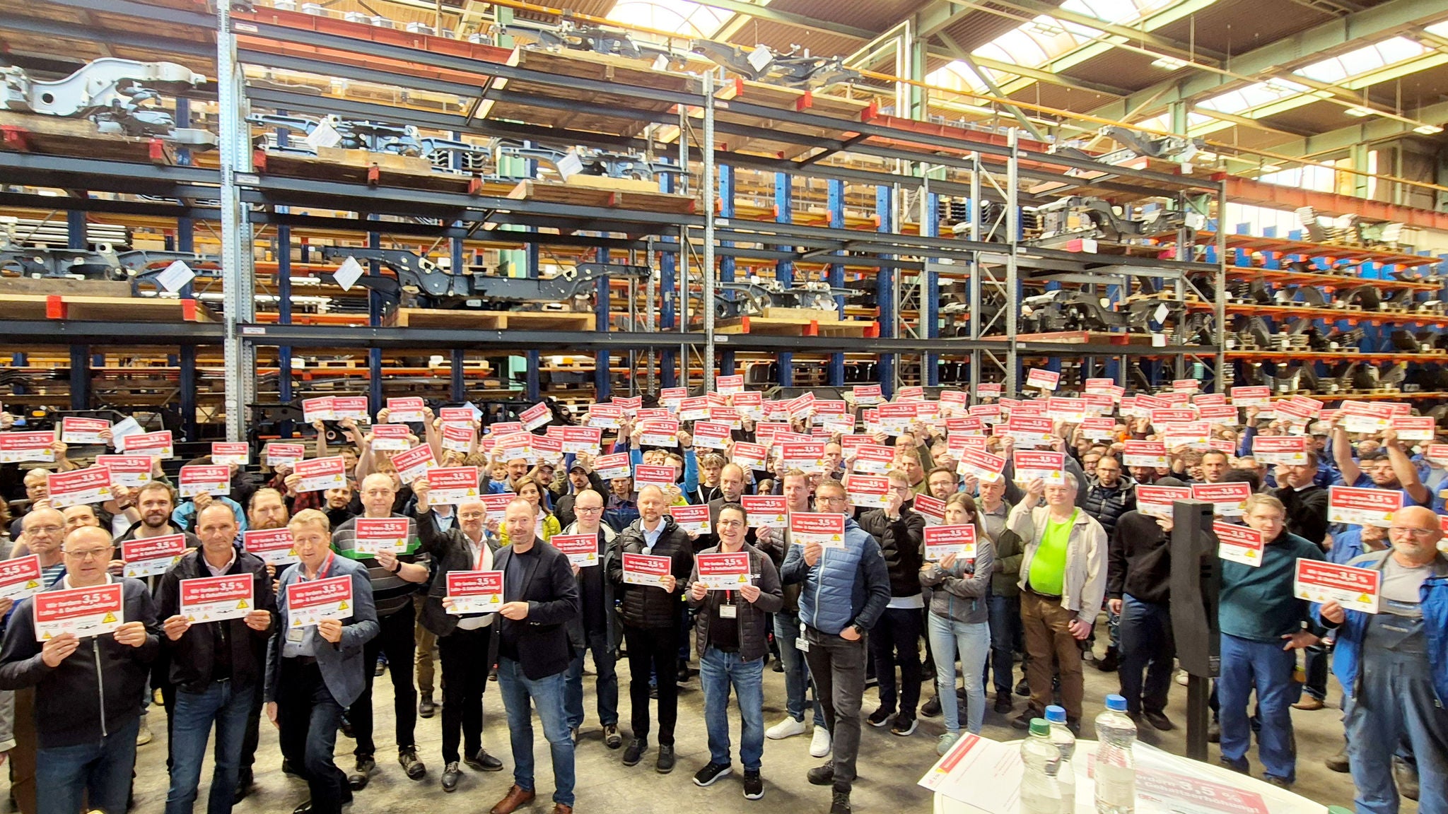 Eine große Gruppe von Beschäftigte bei der Betriebsversammlung in einer Lagerhalle mit Schildern "Wir fordern 3,5 Prozent"