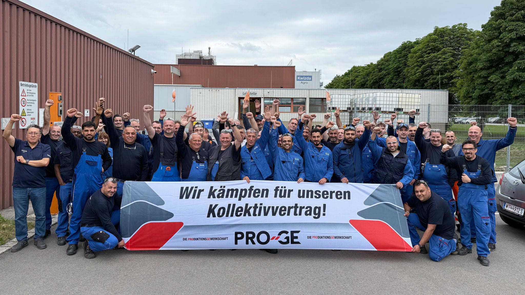 Gruppenfoto der Belegschaft vor einem Werksgebäude mit einem Transparent "Wir kämpfen für unseren Kollektivvertrag!"