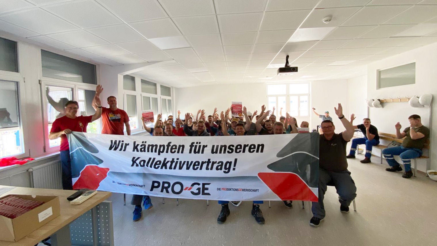 Teilnehmer:innen der Versammlung, sitzend, mit Transparent "Wir kämpfen für unseren Kollektivvertrag!"
