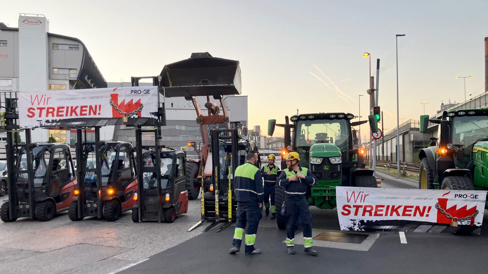 4 Arbeiter mit 2 Traktoren, 1 Bagger und 3 Gabelsaplern bei Sonnenaufgang auf dem Werksgelände, Transparente "Wir streiken!" auf den Fahrzeugen montiert