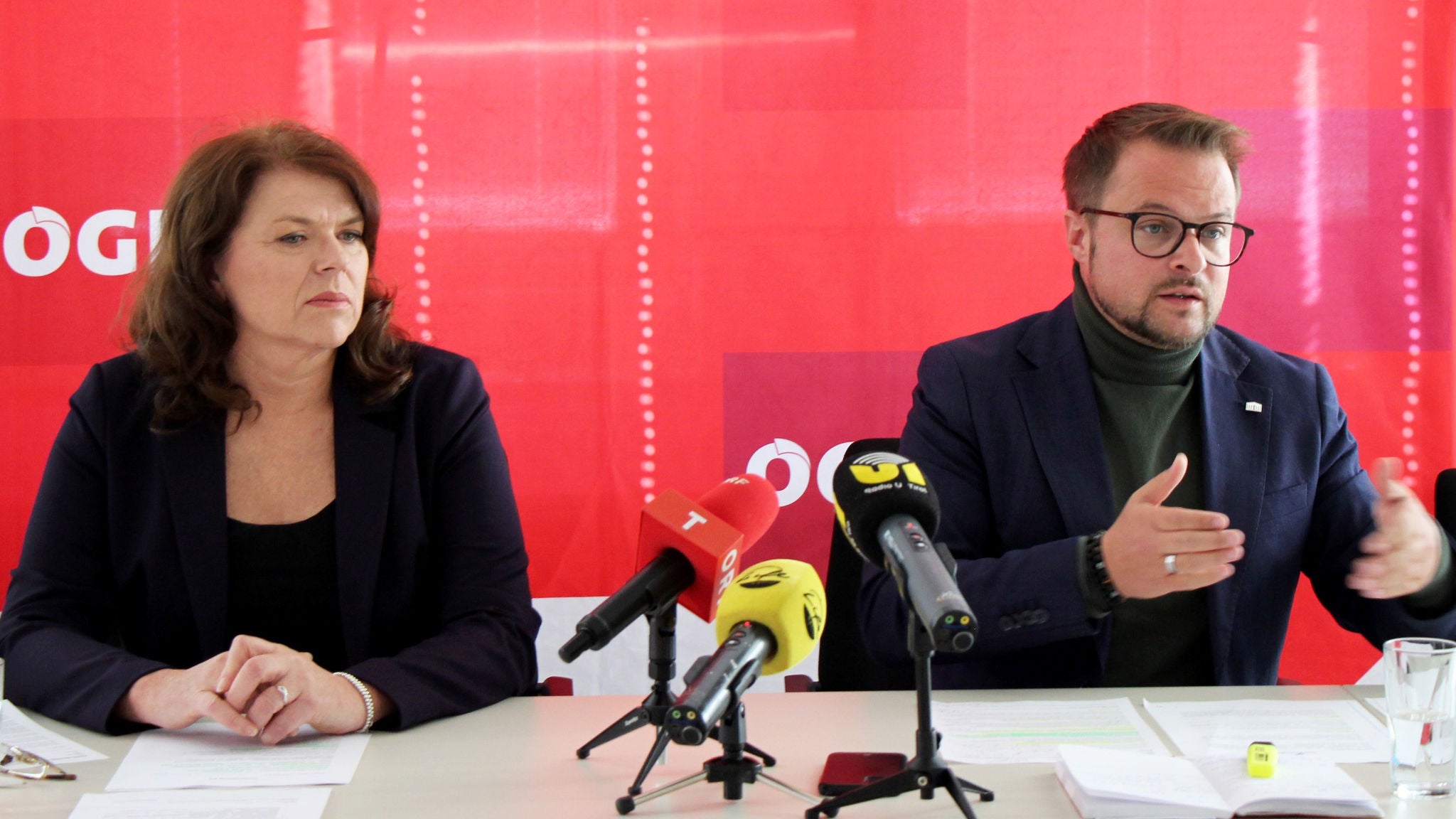 Zwei Personen an einem Tisch vor rotem Hintergrund mit ÖGB-Logos, vor ihnen drei Mirkofone
