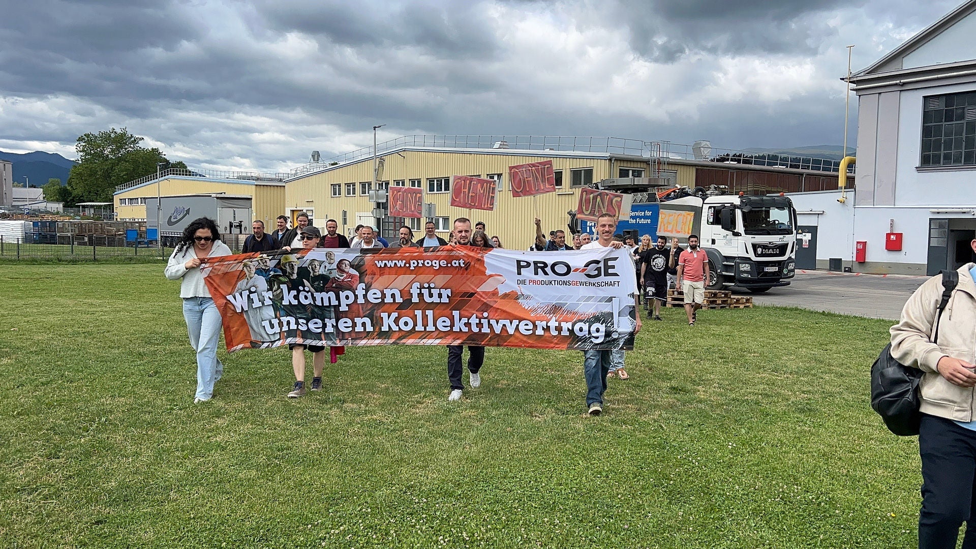Beschäftigte ziehen mit einem Transparent "Wir kämpfen für unseren Kollektivvertrag!" und Schildern "Keine Chemie ohne uns" über eine Wiese vor dem Werksgelände