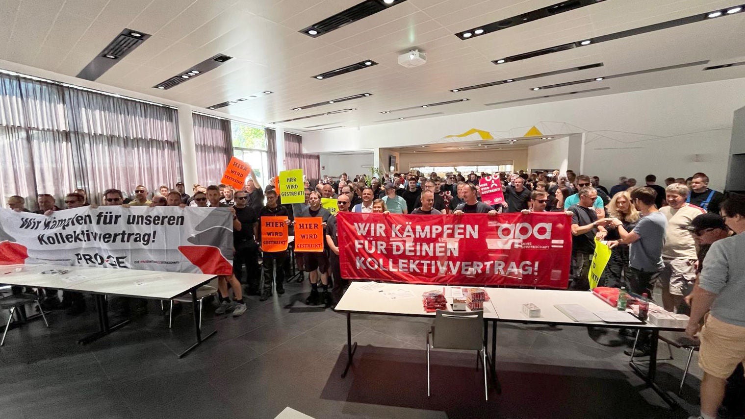 Teilnehmer:innen der Versammlung, sitzend, mit weißem Transparent "Wir kämpfen für unseren Kollektivvertrag! PRO-GE" und rotem Transparent "Wir kämpfen für deinen Kollektivvertrag! GPA"