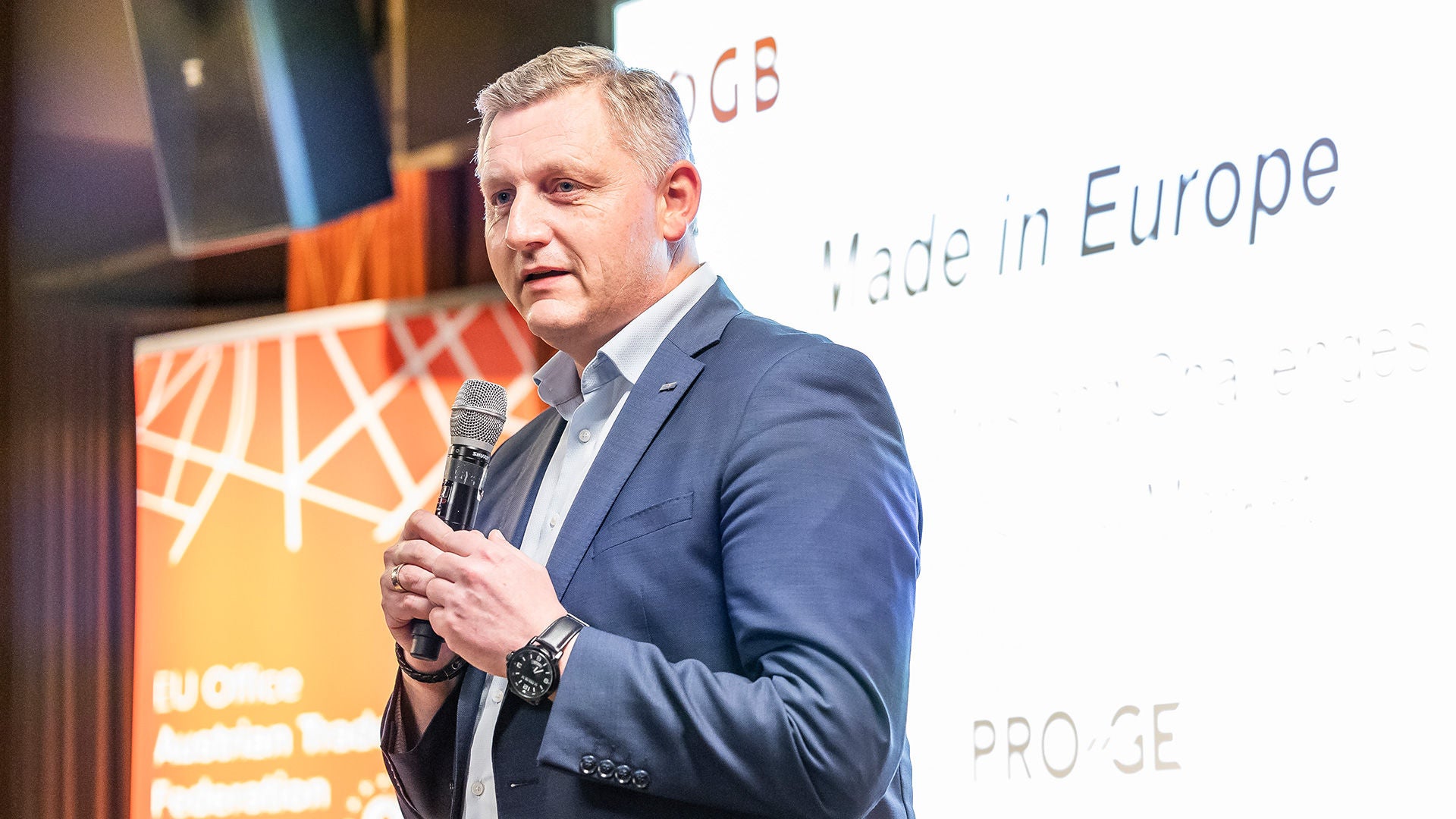 Reinhold Binder mit Mikrophon vor einem Screen mit ÖGB-Logo und Schriftzug "Made in Euope"