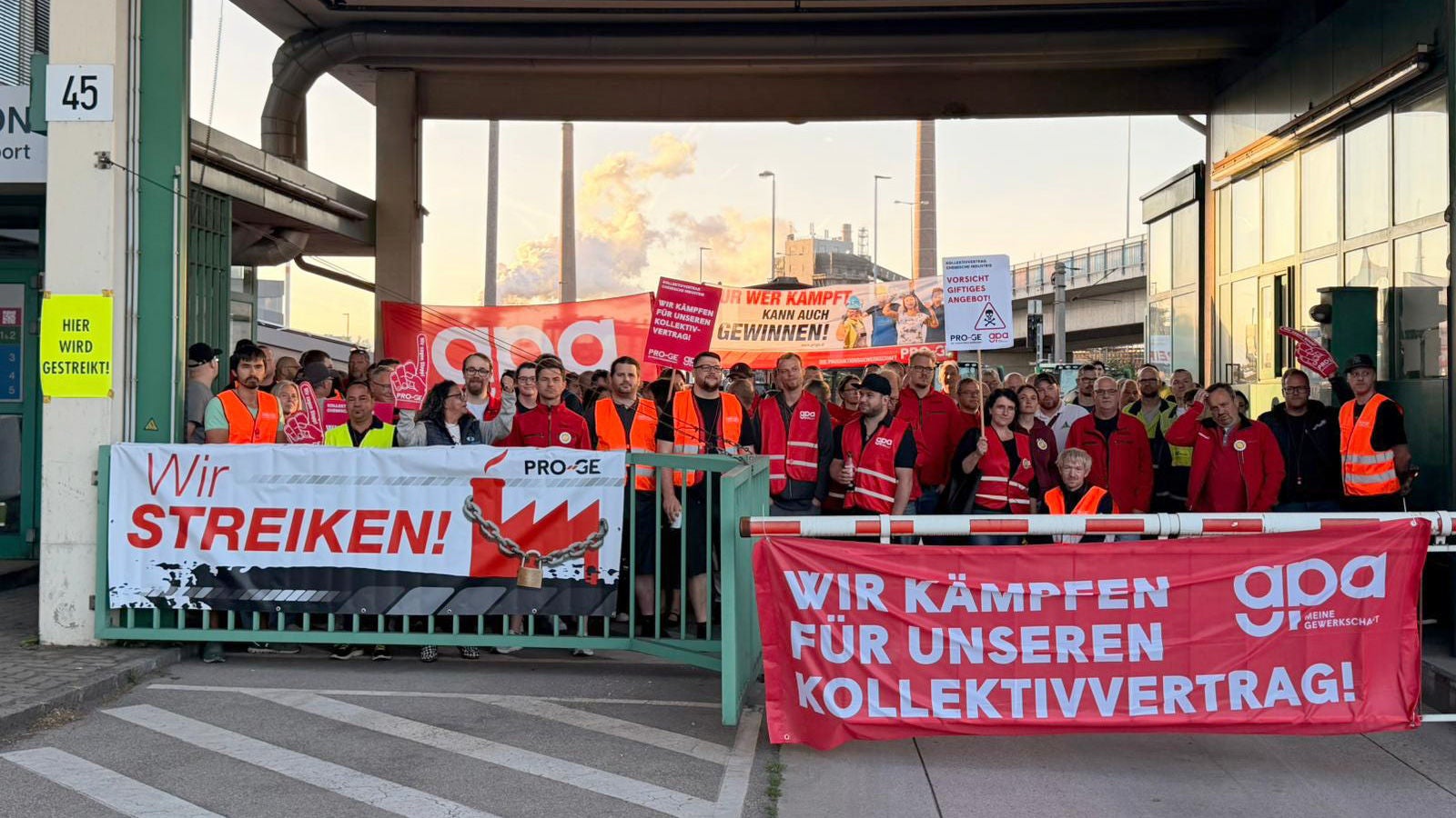Eine Gruppe Streikender an einer Werkseinfahrt mit Transparenten "Wir streiken!" und "Wir kämpfen für unseren Kollektivvertrag!"