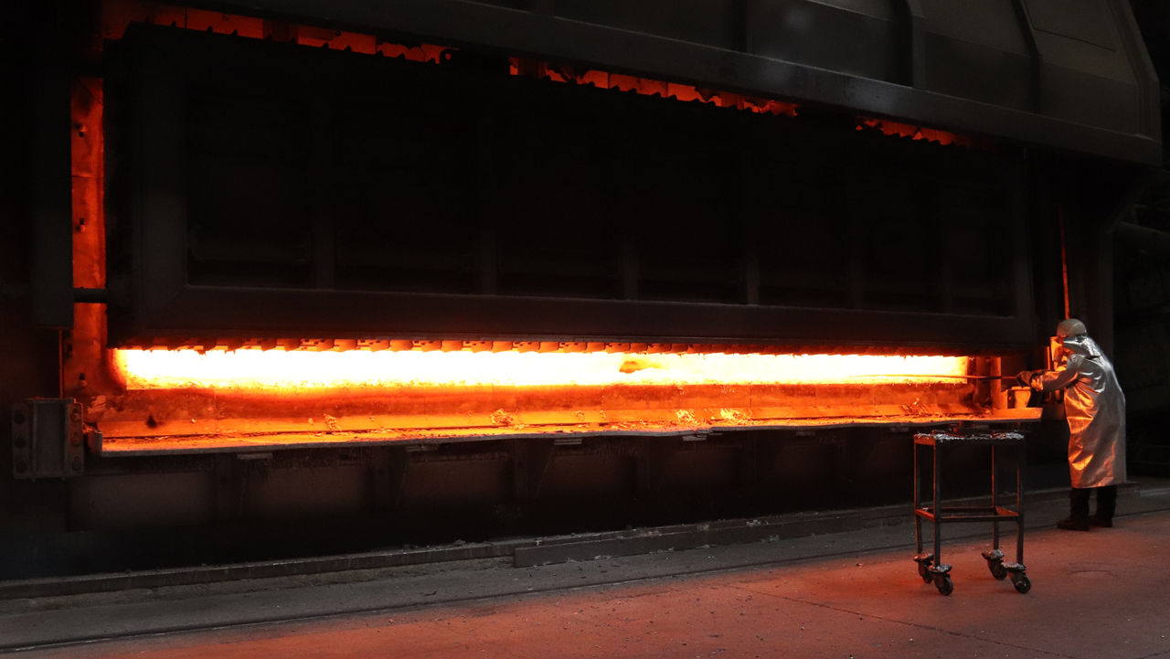 Man sieht die Silhouette eines Arbeiters vor den Flammen eines Hochofens, der eine Probe entnimmt.